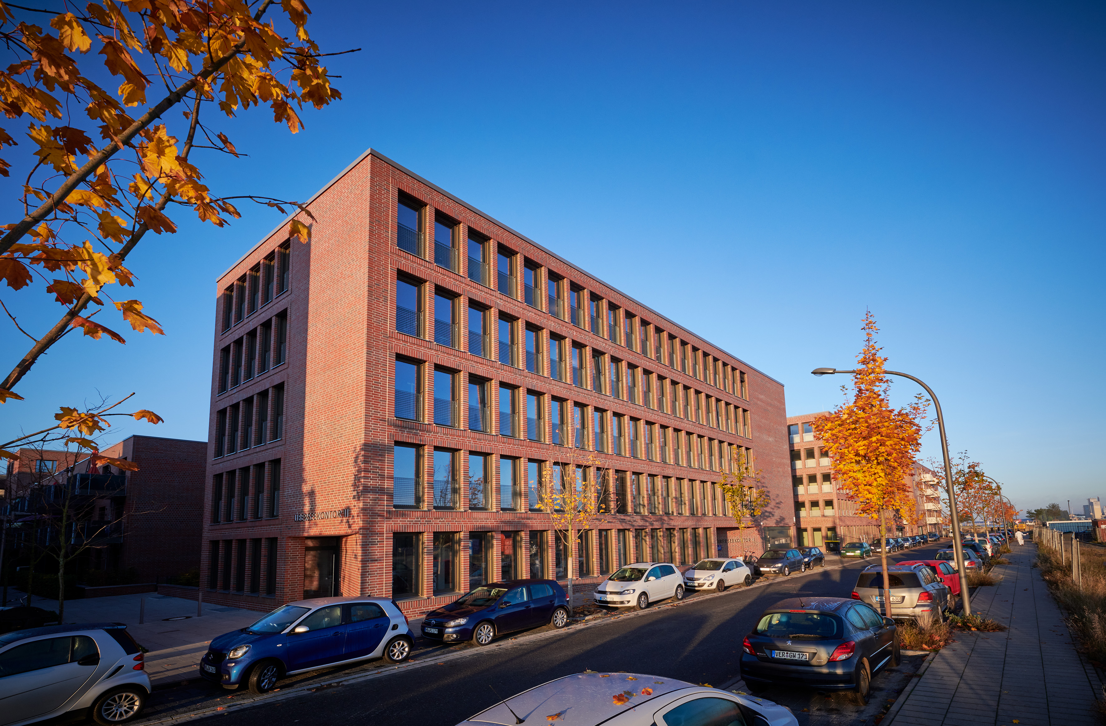 Modernes Bürogebäude aus rotem Backstein an einer Straße mit parkenden Autos und herbstlichen Bäumen.