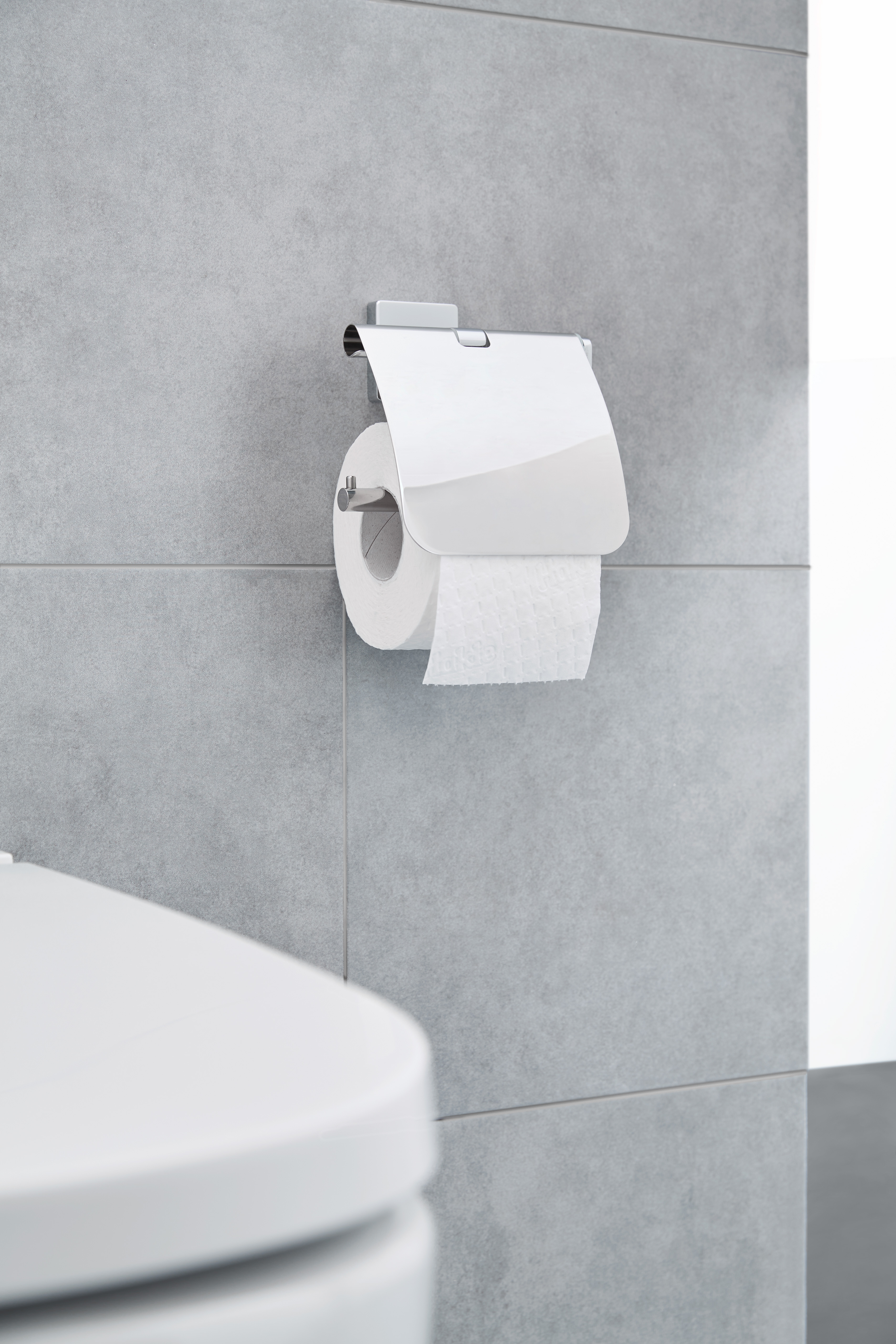 Moderner Toilettenpapierhalter aus Edelstahl an einer grauen Fliesenwand, neben einer weißen Toilette.