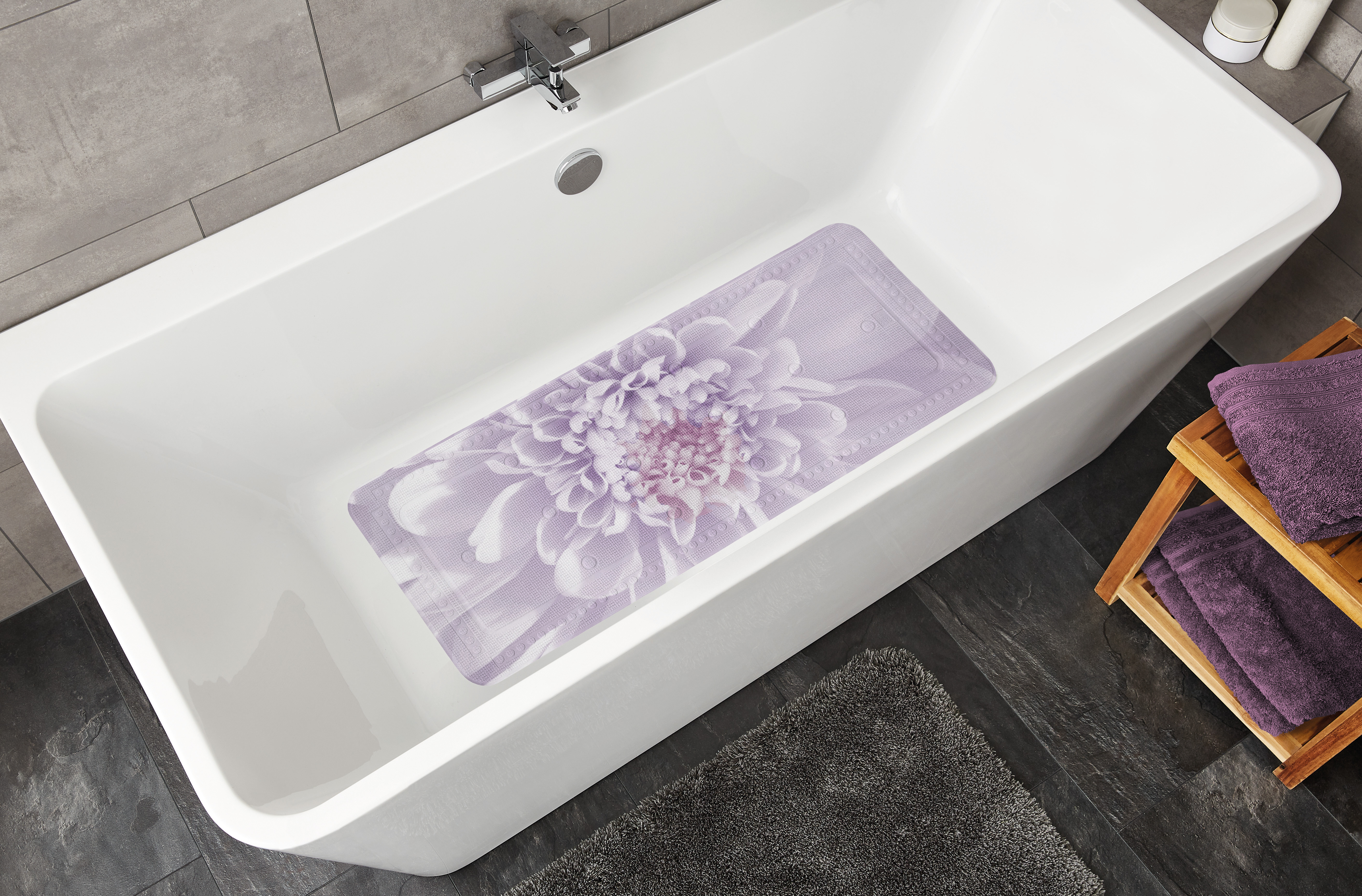 Lila Badematte mit Blumenmuster in einer modernen weißen Badewanne, umgeben von grauen Fliesen und Handtüchern.